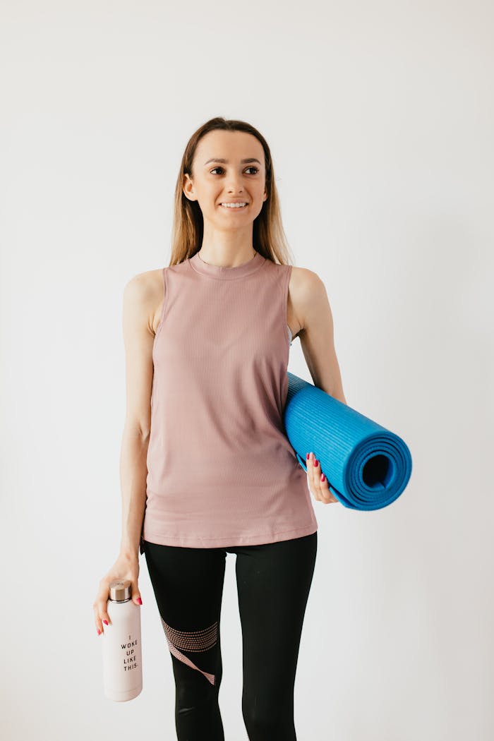 about-01 Positive young female athlete in sportswear standing isolated against white wall with blue yoga mat and water bottle while looking away with smile